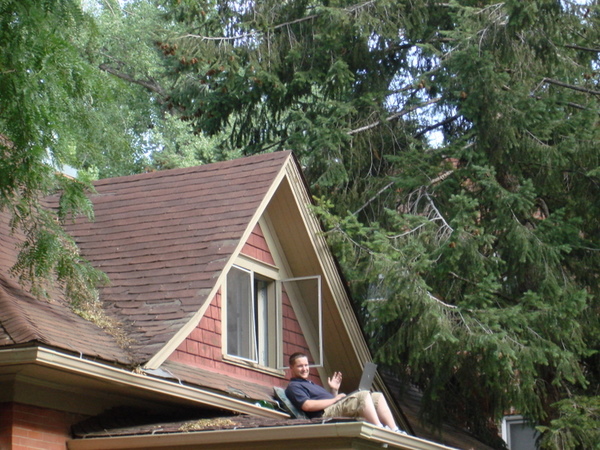 A guy with a laptop on the roof of his house waves.