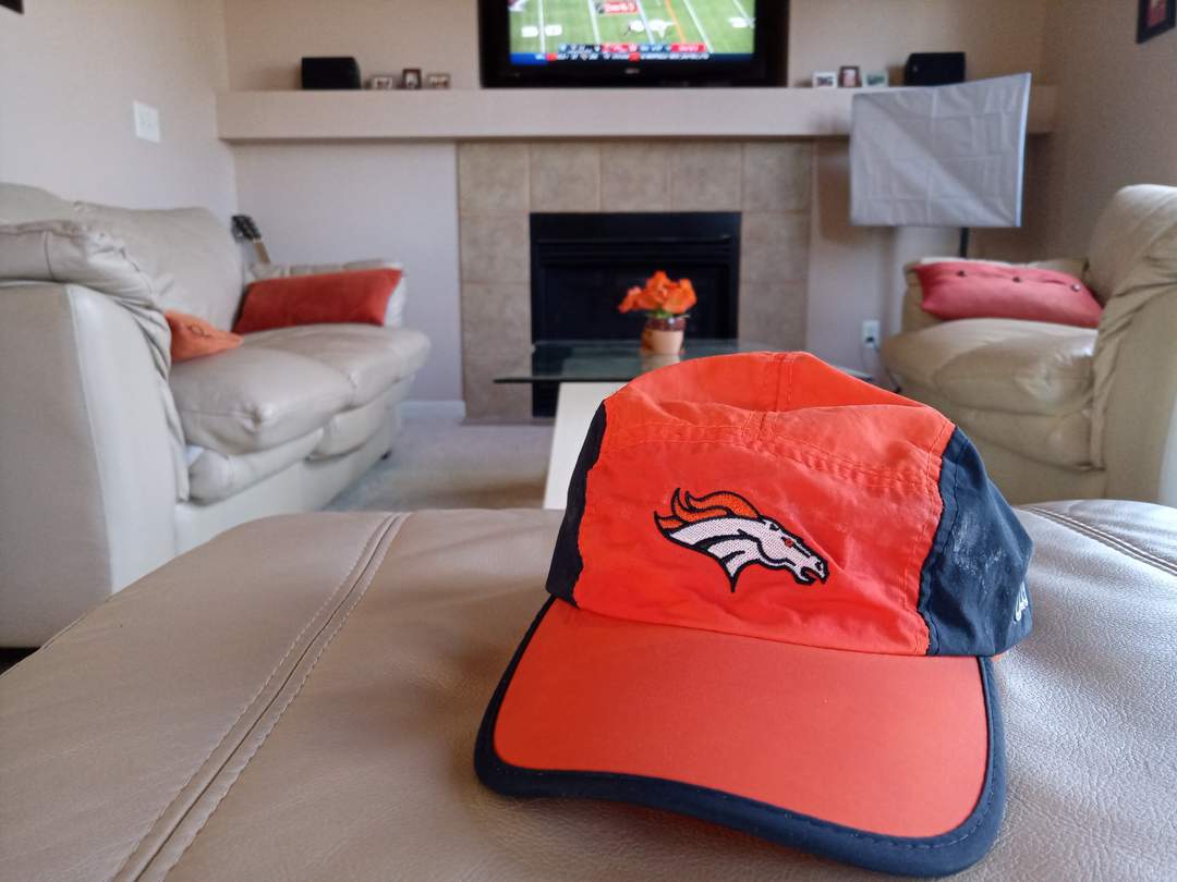 orange and blue Denver Broncos cap on beige ottoman in living room with football game on TV