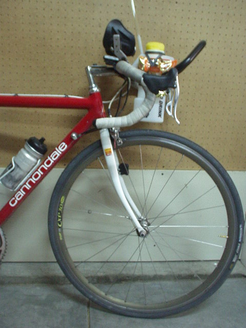front half of red Cannondale road bike with white fork and aerobars containing water bottle against pegboard