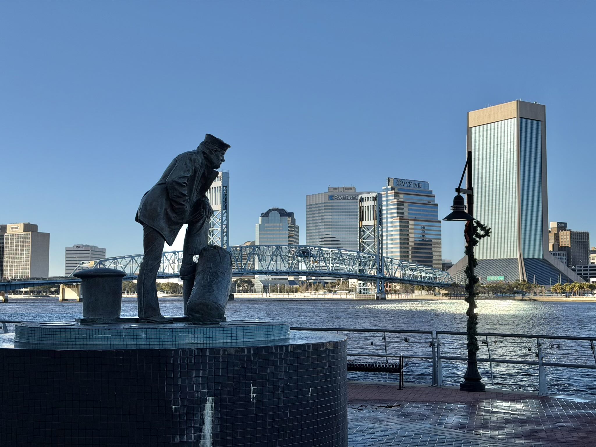 The Lone Sailor Statue on the Southbank Riverbank, with tall office buildings on the other side of the St. Johns River in downtown Jacksonville.