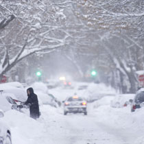 car driving down snow-covered street while a woman in black coat digs out car