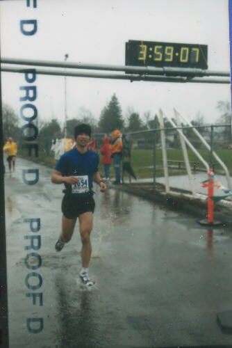 Felix Wong wearing blue jersey and black beanie, crossing rain-soaked finish line in 3:59:07