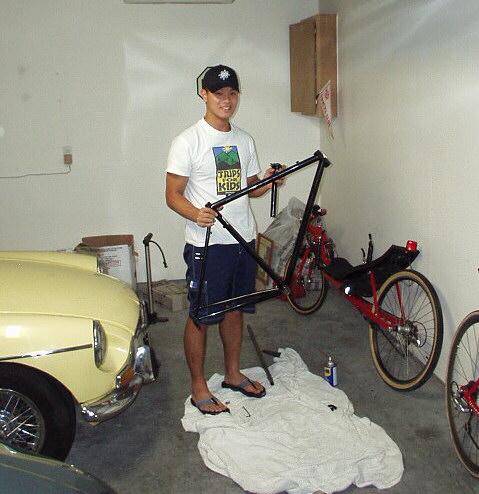 yellow MGB, David Hung in white shirt holding a black road bike frame and standing on white towel, red recumbent leaned against white garage wall