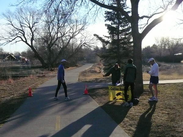 Jonathan Zeif on Spring Creek Trail, two orange cones, three people standing next to yellow Fort Collins Running Club sign