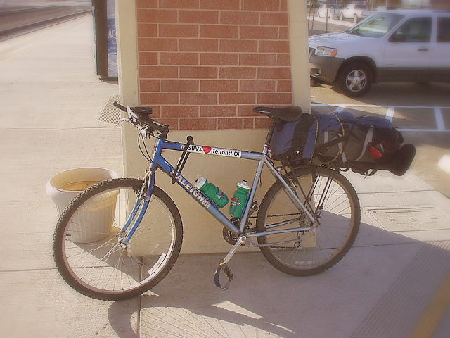 blue Raleigh M500 mountain bike with a bumper sticker that says SUVs [heart] terrorist oil and a large blue camping backpack strapped to the rear rack