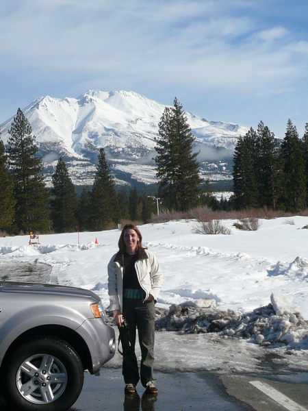 red-haired woman wearing white jacket next to silver Ford Escpe in front of a snow-covered field and Mount Shasta in the background.