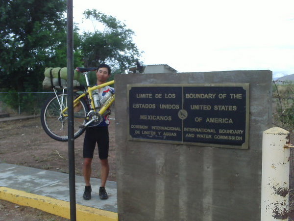 Felix Wong holding up a yellow mountain bike at chest level next to a sign that says Boundary of the United States of America
