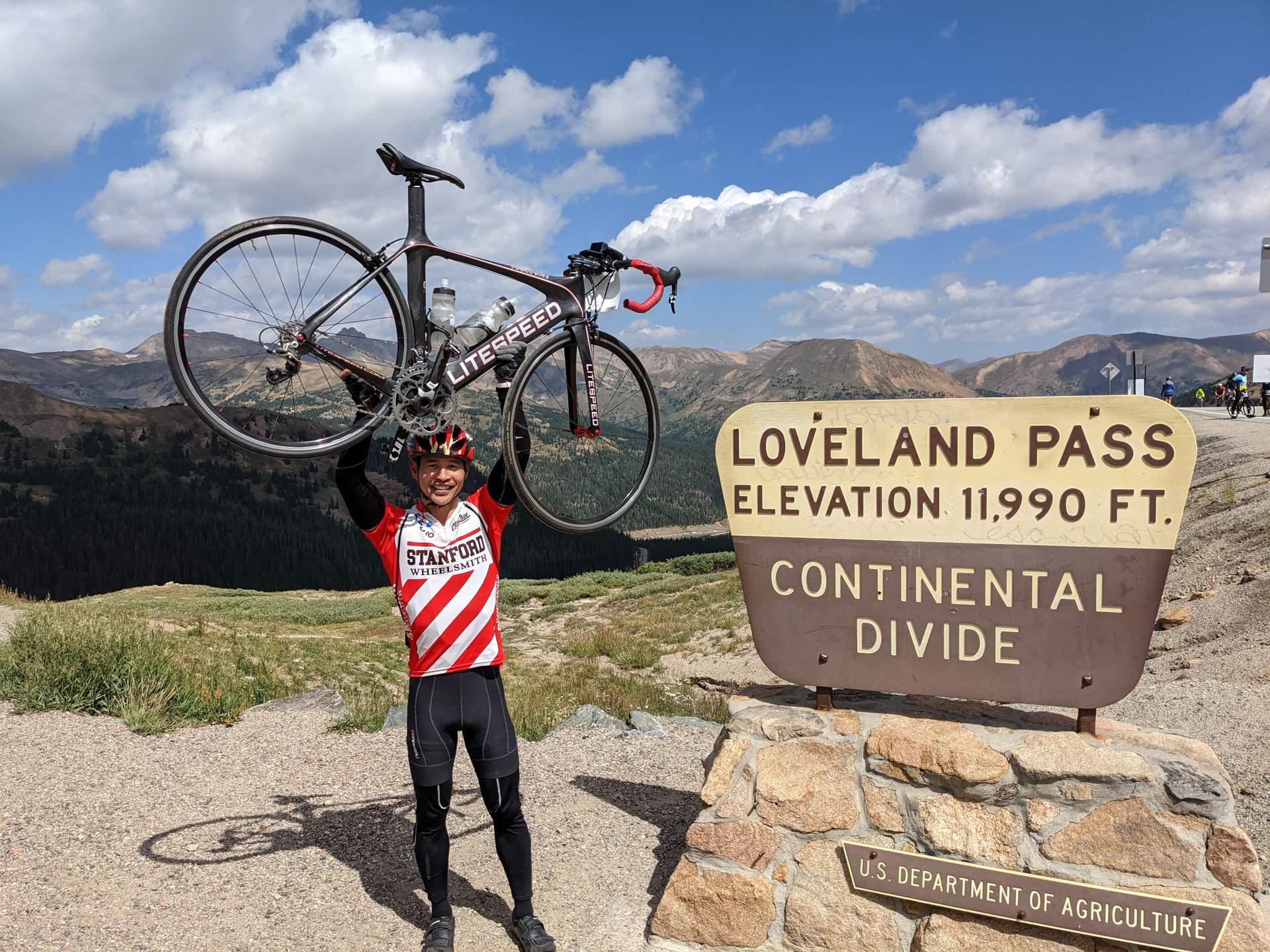 Felix Wong wearing a red and white Stanford jersey lifting his black Litespeed Archon C2 road bike over his head at Loveland Pass, Elevation 11990 feet.