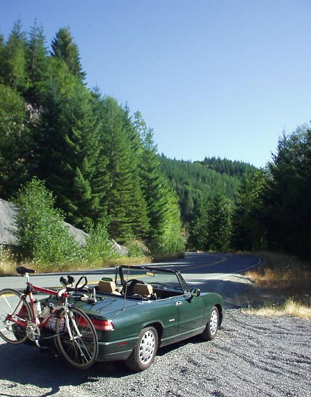 green 1991 Alfa Romeo Spider Veloce, with top down, carrying a red Cannondale road bike, on gravel shoulder next to a road going towards Mount Saint Helens.