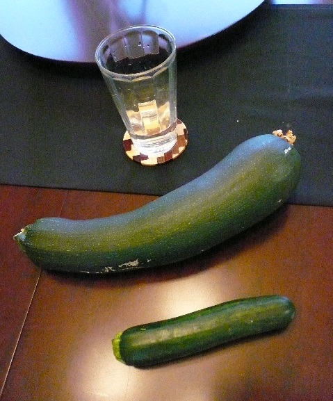 A big zucchini next to a small zucchini on a wooden table next to a glass.