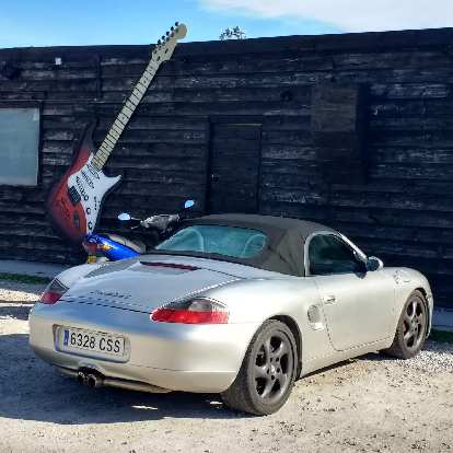 A silver, early 2000s Porsche Boxster next to a blue motorbike and a red and white electric guitar.
