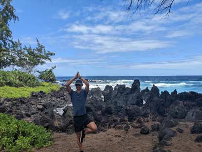 Felix doing tree pose in front of some rocks and the ocean at Laupahoehoe.