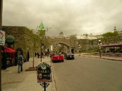 In the historic center of Quebec City. valet parking sign, person walking on sidewalk, red and black car in front of brick archway and green tower