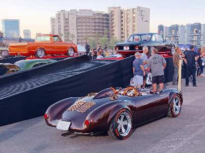 A Porsche 550 Spyder in merlot, with an orange truck and black coupe in the background.