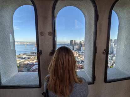 Andrea peering out windows to east from the top of Coit Tower, including a view of the Bay Bridge.