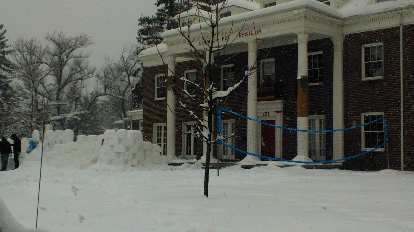 Some folks were building a wall out of snow bricks by this snowman in front of this fraternity.