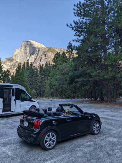 The black John Cooper Works edition MINI Convertible by a white recreation vehicle in a parking lot at Curry Village, with the west face of Half Dome towering above.