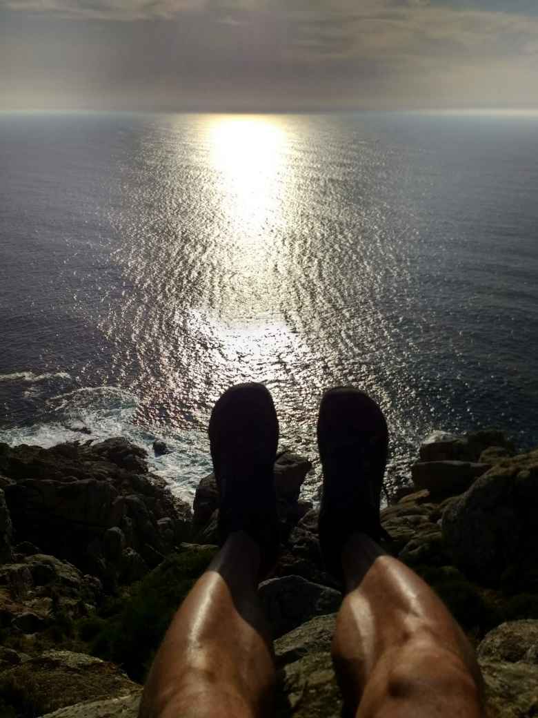 Felix Wong's legs and feet overhanging a rocky cliff on the western shoreline of Fisterra, sun reflecting off the Atlantic Ocean