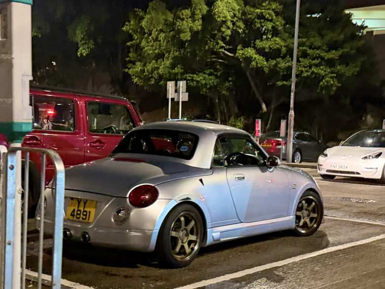 A silver Daihatsu Copen (2002-2012) with aftermarket 6-spoke wheels and a dual-exit exhaust.