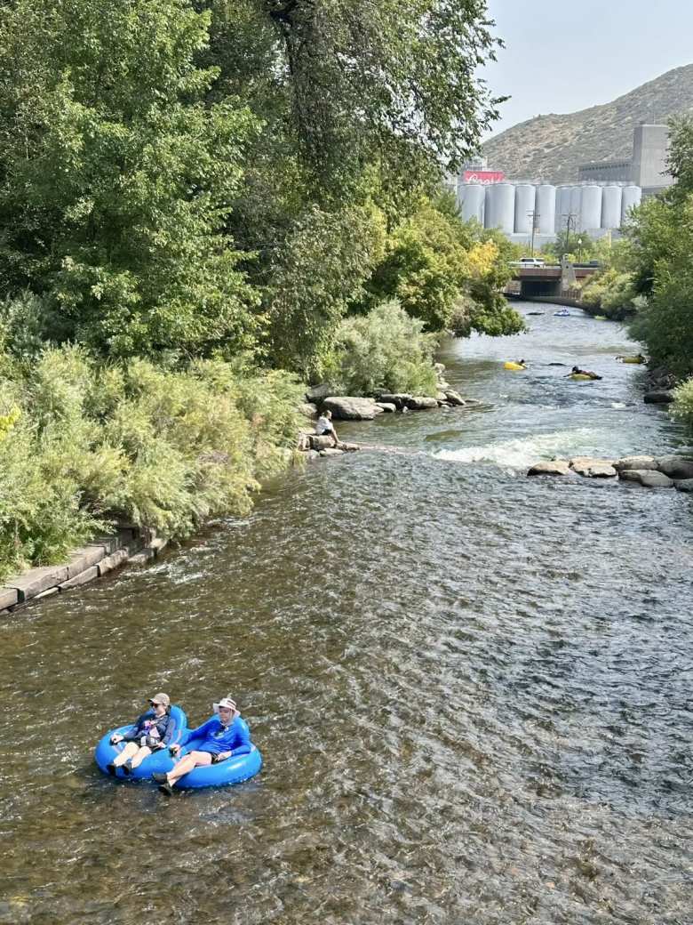 Two people floating down Clear Creek in downtown Golden, with the Coor's factory in the background.