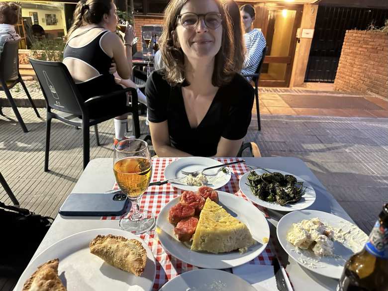 Eli sitting at a table with empanandas, tortilla, tostada con tomate, pimentos de Padrón, and ensalada rusa.