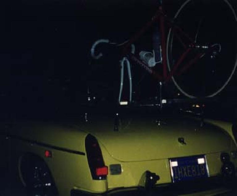 red bicycle mounted on top of yellow MGB roadster at night.