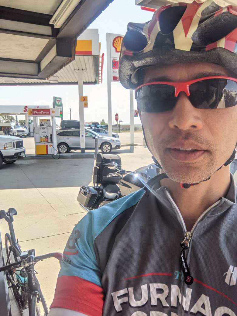 Felix Wong wearing grey/turquoise/red Furnace Creek 508 bicycle jersey, red sun glasses, with carbon fiber bicycle and cruiser motorcycle outside Shell gas station