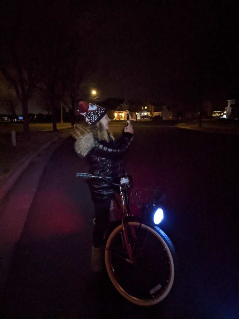 Andrea standing next to a Huffy Cranbrook cruiser bike with a bright headlight, taking a picture of neighborhood Christmas lights at night.
