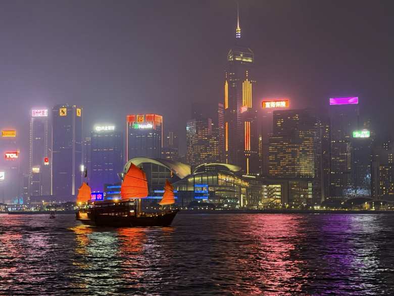 A traditional red-sailed junk boat (available for rent) glides across Victoria Harbour against the shimmering backdrop of the Hong Kong skyline at night.