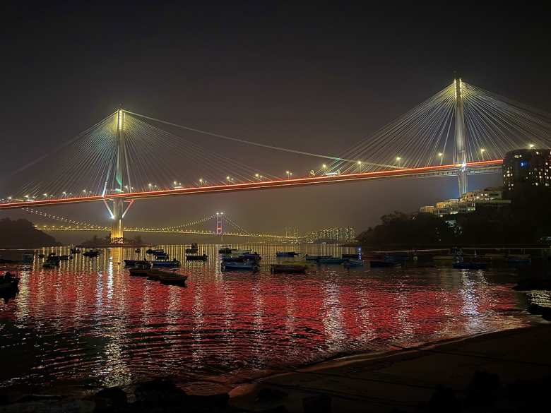 The illuminated Ting Kau Bridge stretches across the water, creating a brilliant display of light and reflection in the evening.