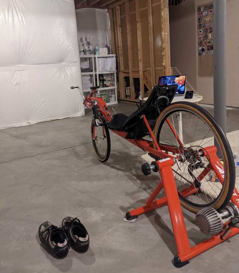My Porsche Guards Red Reynolds Wishbone recumbent, Performance Scattante cycling shoes, iPad Pro, and iPhone 14 Pro in the basement.