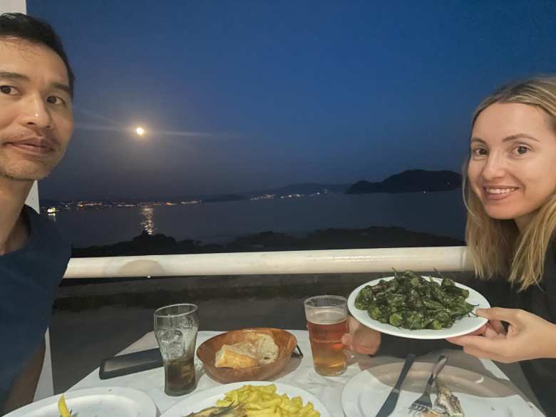 Felix and Andrea eating a dinner of fried peppers, fish, french fries, bread, and Estrella Galicia at Restaurante Rodas, with a full moon and the Atlantic Ocean behind.