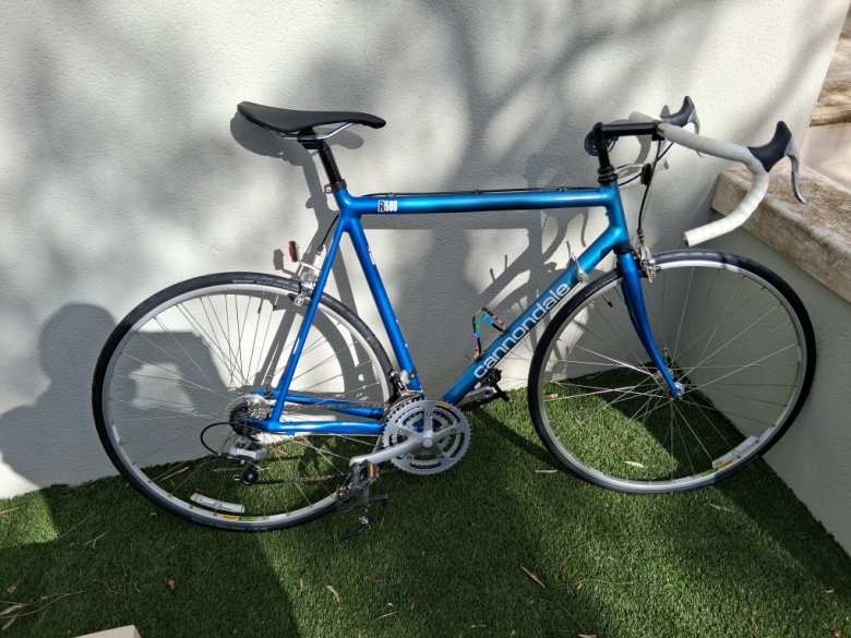 a blue 1993 Cannondale R500 leaning against a white wall on artificial turf