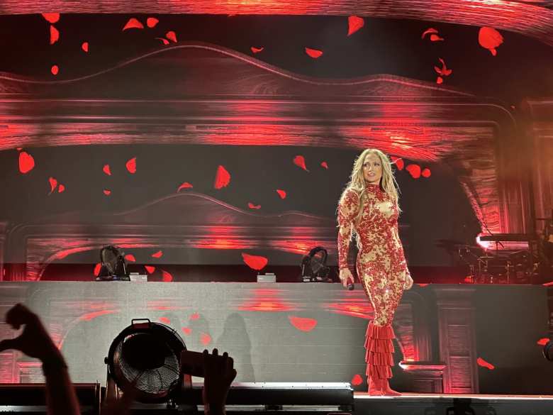 Jennifer Lopez performing live in a red and gold floral-patterned dress with ruffled sleeves and matching red boots.