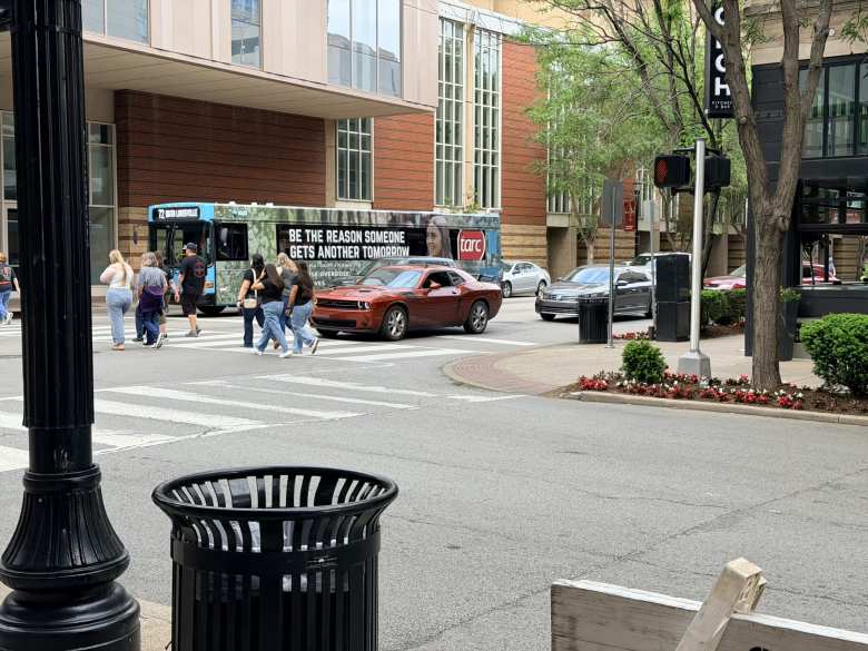 Pedestrians walk by a bronze Dodge Challenger in downtown Louisville.
