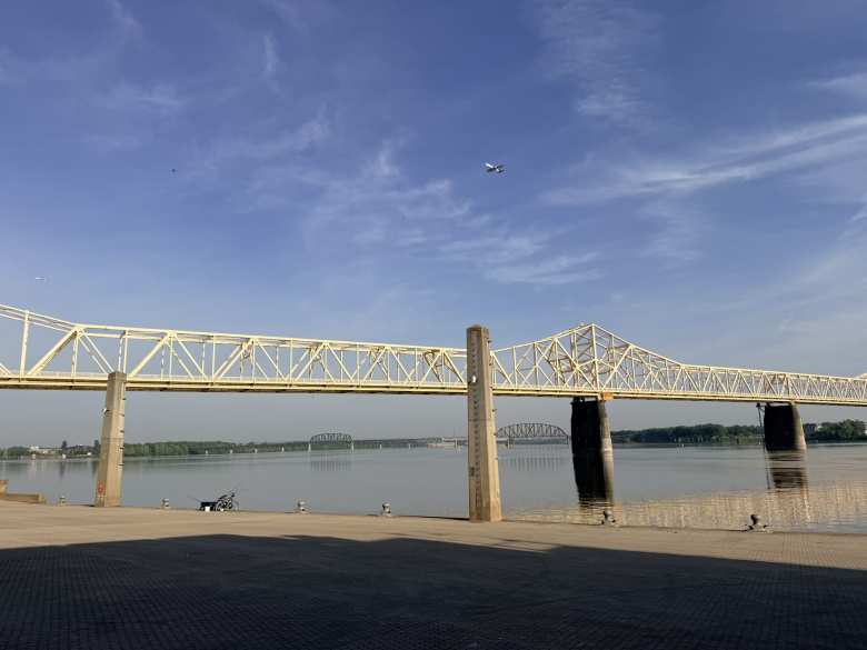 A plane flies over the Ohio River and the George Rogers Clark Memorial Bridge. Someone set up some fishing poles at the base of the bridge on the waterfront's esplanade.