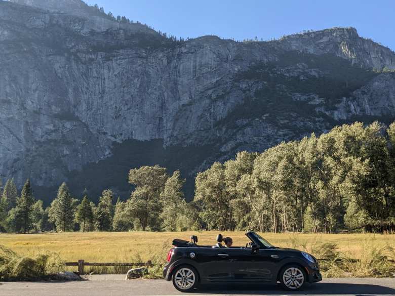 A side profile of the black MINI John Cooper Works Convertible in Yosemite, with a field and trees behind and a tall granite wall towering over all of them.