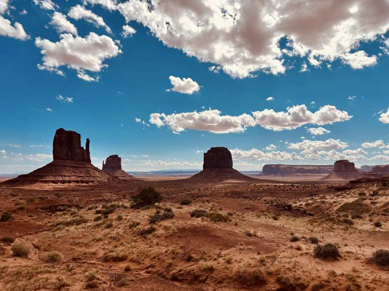 Sunlight and shadows dance across the vast desert floor of Monument Valley, where the West Mitten, East Mitten, Merrick, and Elephant buttes rise beneath a partly cloudy sky.