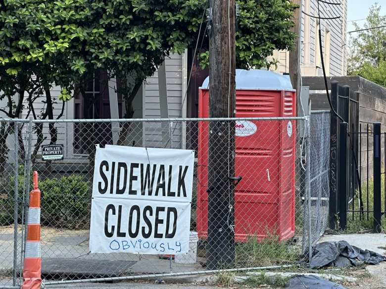 White sign on chainlink fence that originally read "Sidewalk closed," but someone had scrawled "obviously" on it.