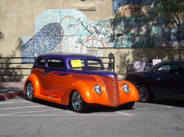 An orange and purple hot rod based off a Ford Sedan of the 1930s, with a nice tile mural behind.
