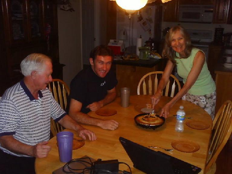 Dick Vail, Tim Vail, Dee Vail at a kitchen table with Dee cutting some apple pie