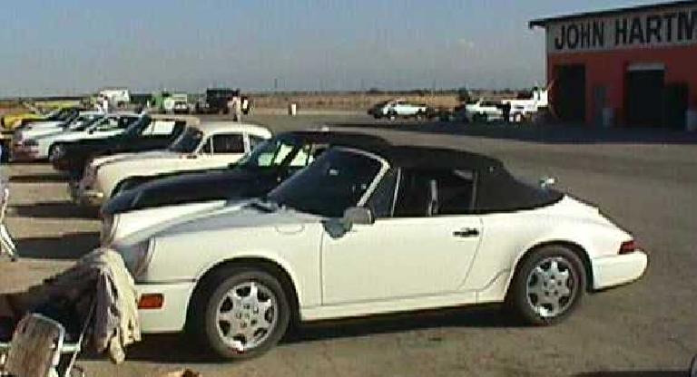 white 1990 Porsche 911 Carrera Cabriolet with black top and a row of Porsches next to it on an airport base