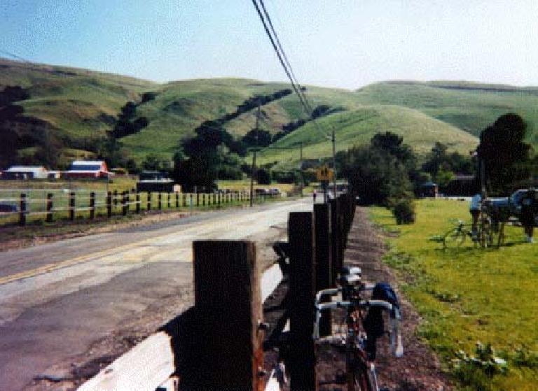 bikes leaned against wooden fence by road, green foothills in background