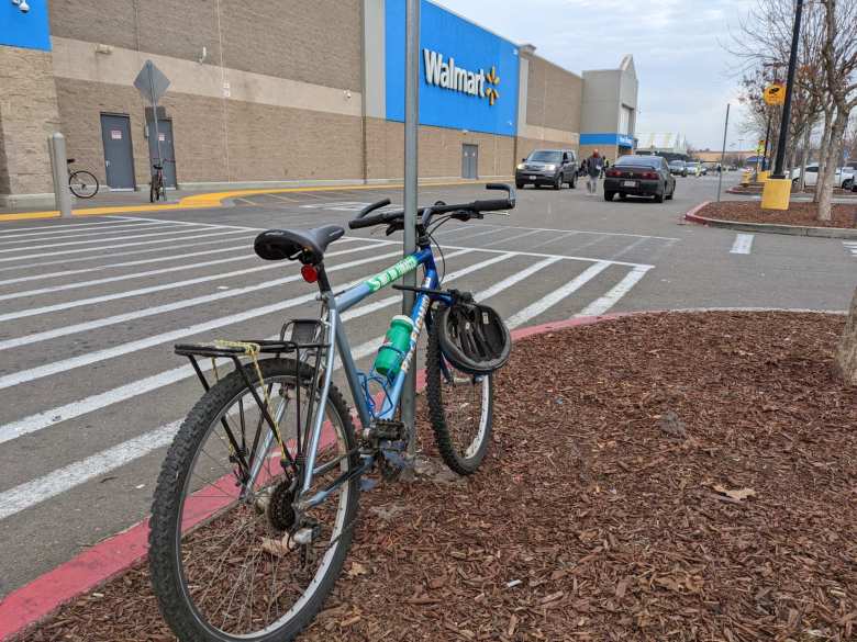 Blue 1996 Raleigh M300 mountain bike with helmet, rear rack, PowerGrips, and a green Gatorade bottle locked to a pole at Walmart in Stockton, California.