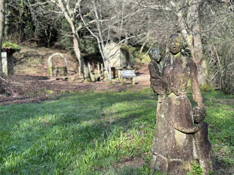 A stone statue of a woman with two kids at the aldea. You can see an hórreo (Galician granary on stilts) in the background.