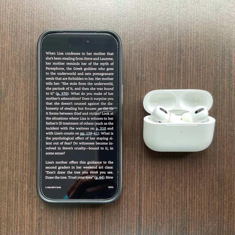 A page from Lisa Brennan-Jobs' "Small Fry" book in the Kindle app on an iPhone 17 Pro next to Apple AirPods Pro earbuds and case.