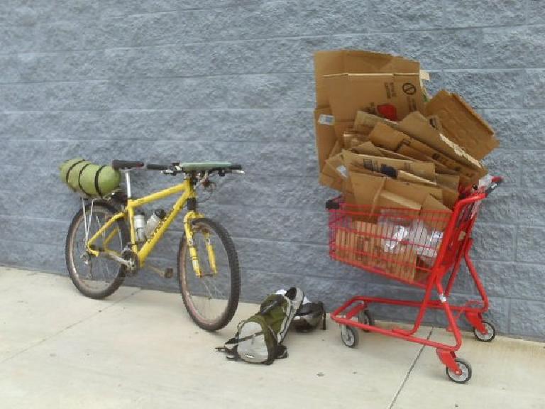 yellow 1996 Cannondale F700 mountain bike with green dry bag strapped to rear rack, green/grey backpack on ground, red shopping card filled with dozens of cardboard boxes