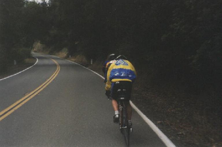 Harry Gretzke wearing yellow and blue Tri-City Triathlon Club windbreaker and riding up a hill behind another cyclist.