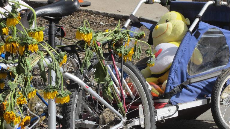 A bicycle adorned with sunflowers and towing a trailer filled with stuffed animals.