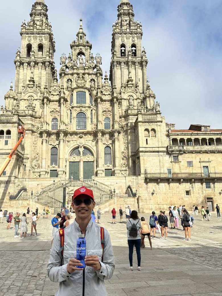 Felix holding a Vaughan bottle and wearing red Carrefour hat in front of the Cathedral of Santiago de Compostela.